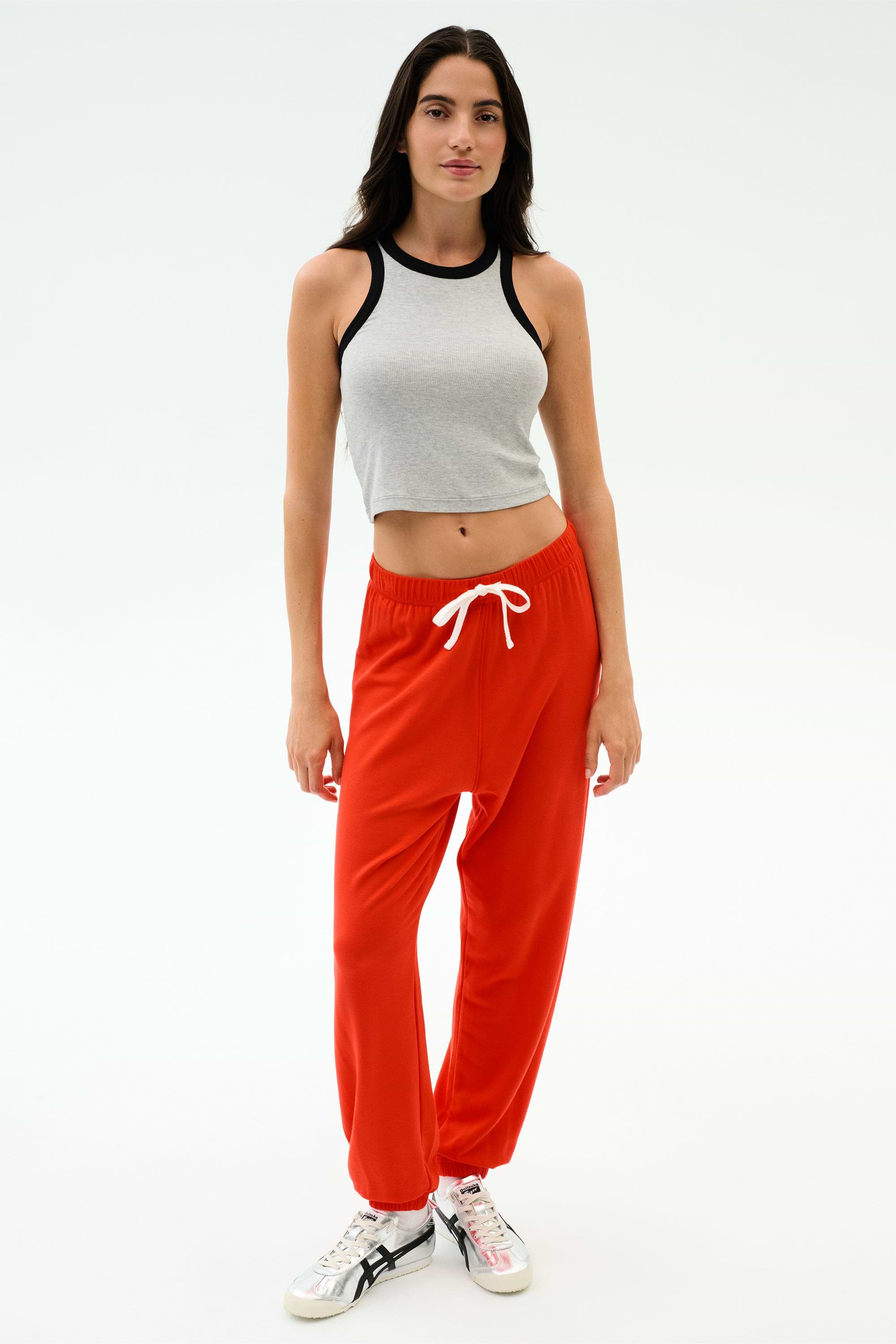 A woman faces forward in a gray sleeveless crop top and SPLITS59 Andie Oversized Fleece Sweatpant in Lotus Red with a white drawstring, paired with white sneakers, set against a plain white background.