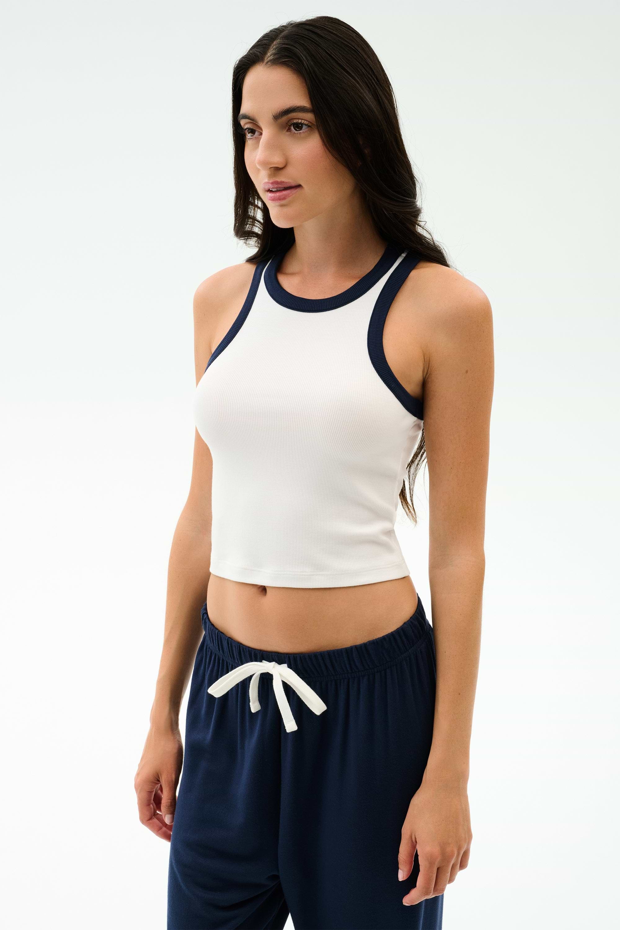 A woman wears the SPLITS59 Kiki Rib Crop Tank in White/Indigo, featuring a baby rib fabric and indigo trim—ideal for gym sessions or casual looks—while standing against a plain light background.