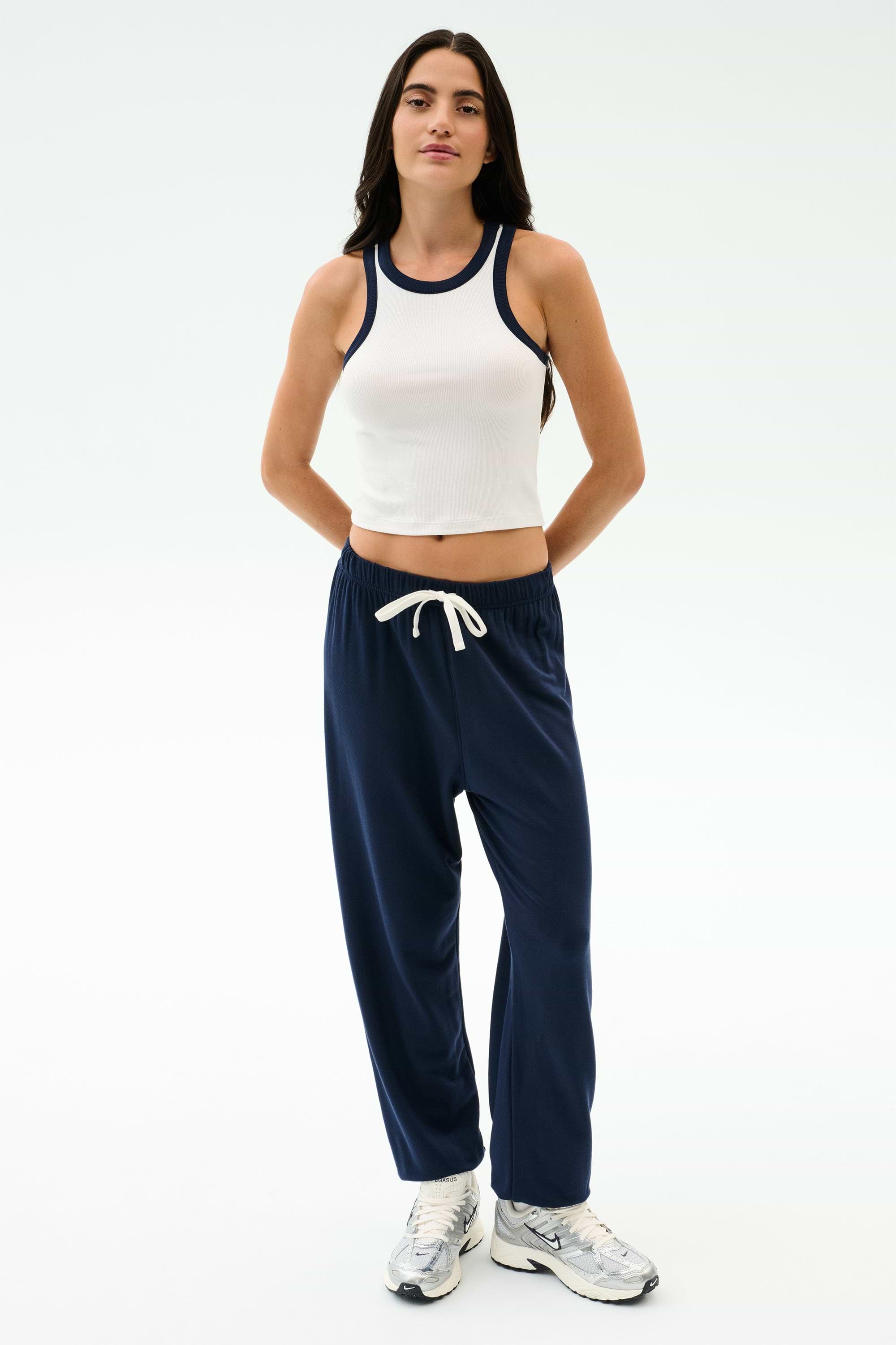 A person stands against a plain background wearing the SPLITS59 Kiki Rib Crop Tank in White/Indigo, navy blue drawstring sweatpants, and white sneakers.
