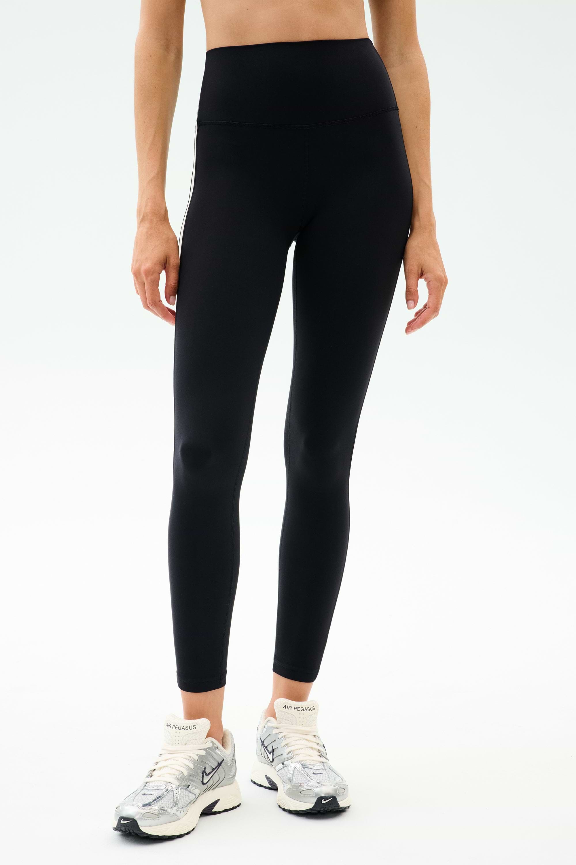 Someone wearing SPLITS59 Clare High Waist Rigor 7/8 leggings in Black/White with white Nike sneakers stands against a plain light background.
