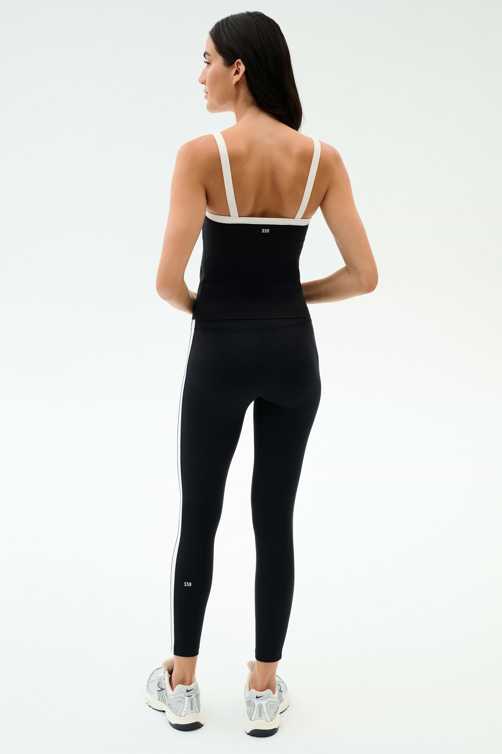 A woman stands facing away, wearing the SPLITS59 Monah Rigor Tank in Black/White with white straps, black side-striped leggings, and white athletic shoes—ideal for gym workouts.