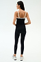 A woman stands facing away, wearing the SPLITS59 Monah Rigor Tank in Black/White with white straps, black side-striped leggings, and white athletic shoes—ideal for gym workouts.