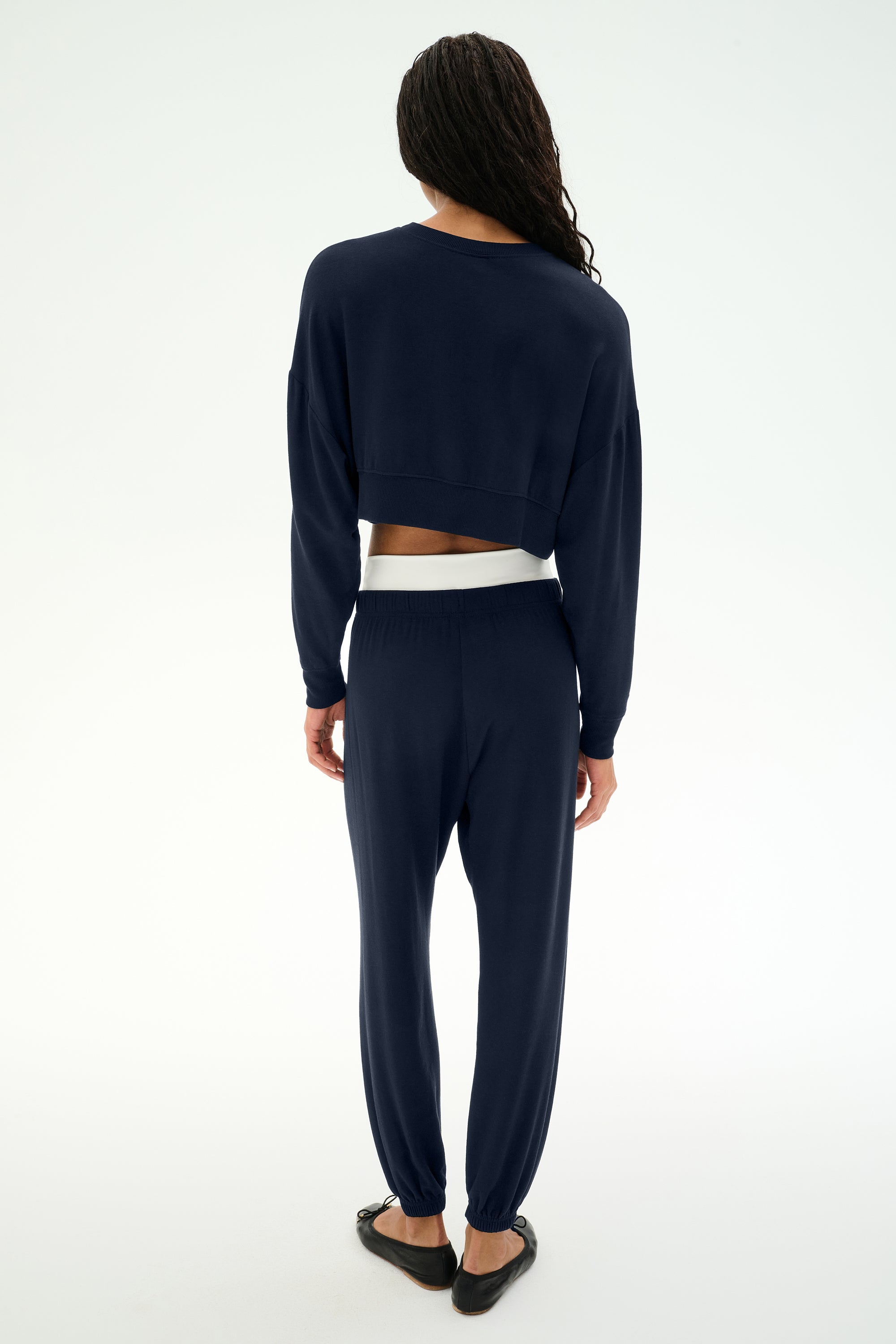 Wearing the SPLITS59 Andie Dual Waist Sweatpant in Indigo/White, this person—proudly made in Los Angeles—stands with their back to the camera, styled with a navy blue long-sleeve top, black flats, and a visible white waistband crafted from ultra-luxe fleece.