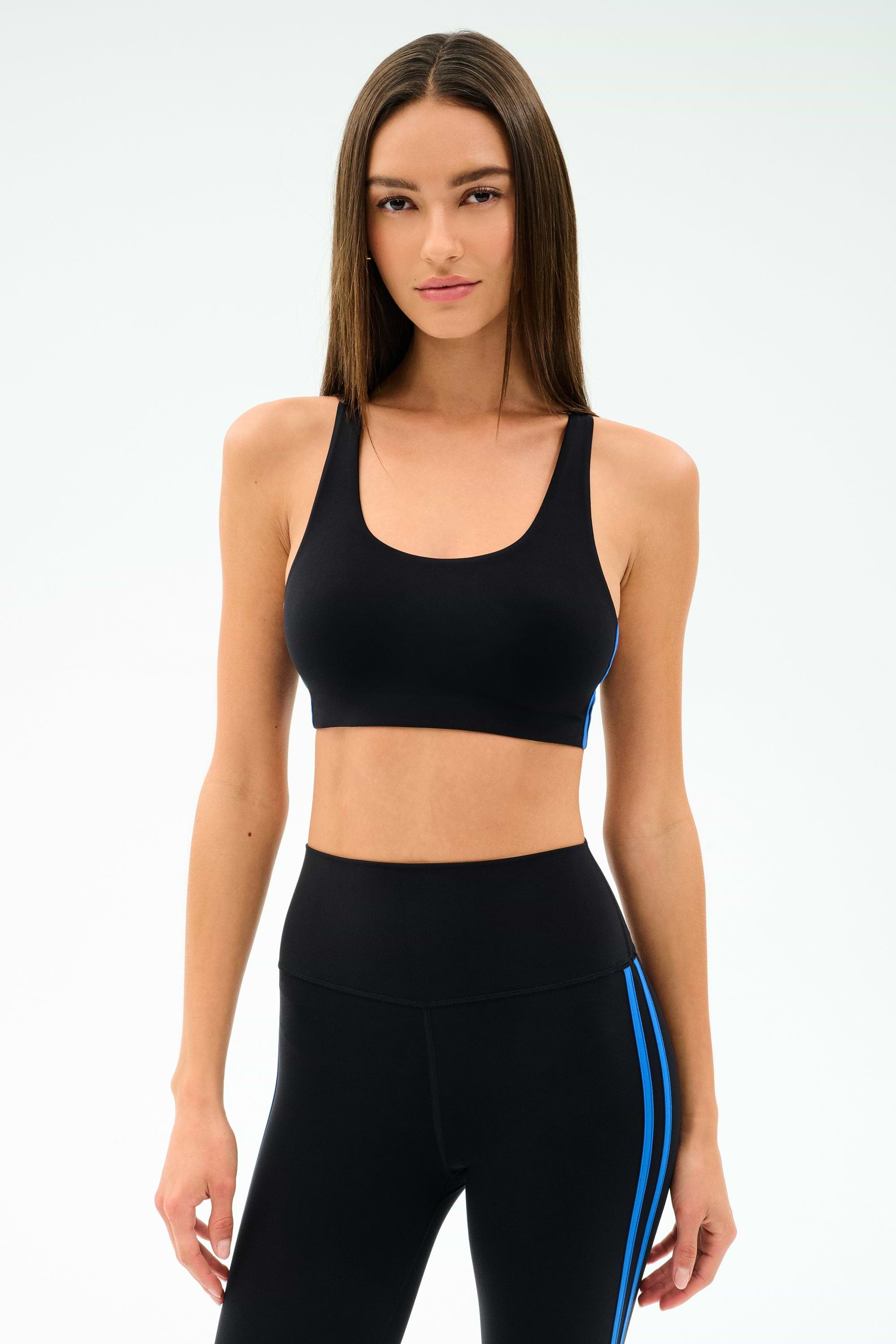 A woman stands against a plain background, wearing SPLITS59’s Ella Airweight Bra in Black/Classic Blue, made from soft nylon spandex for comfort and flexibility, paired with matching black leggings featuring blue side stripes.