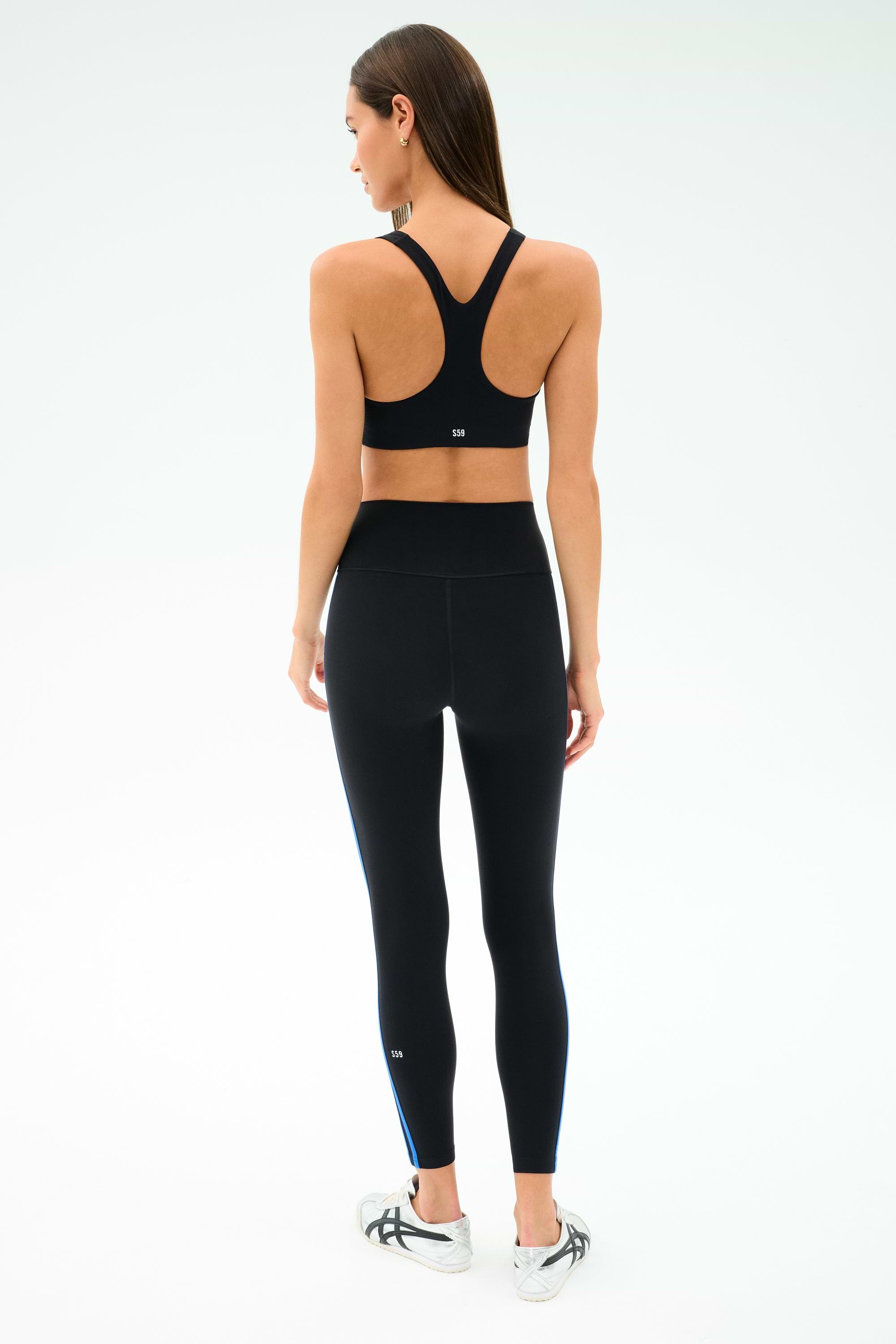A woman faces away, wearing the SPLITS59 Ella Airweight Bra in Black/Classic Blue with high-waisted nylon spandex leggings and white sneakers, set against a plain white background.