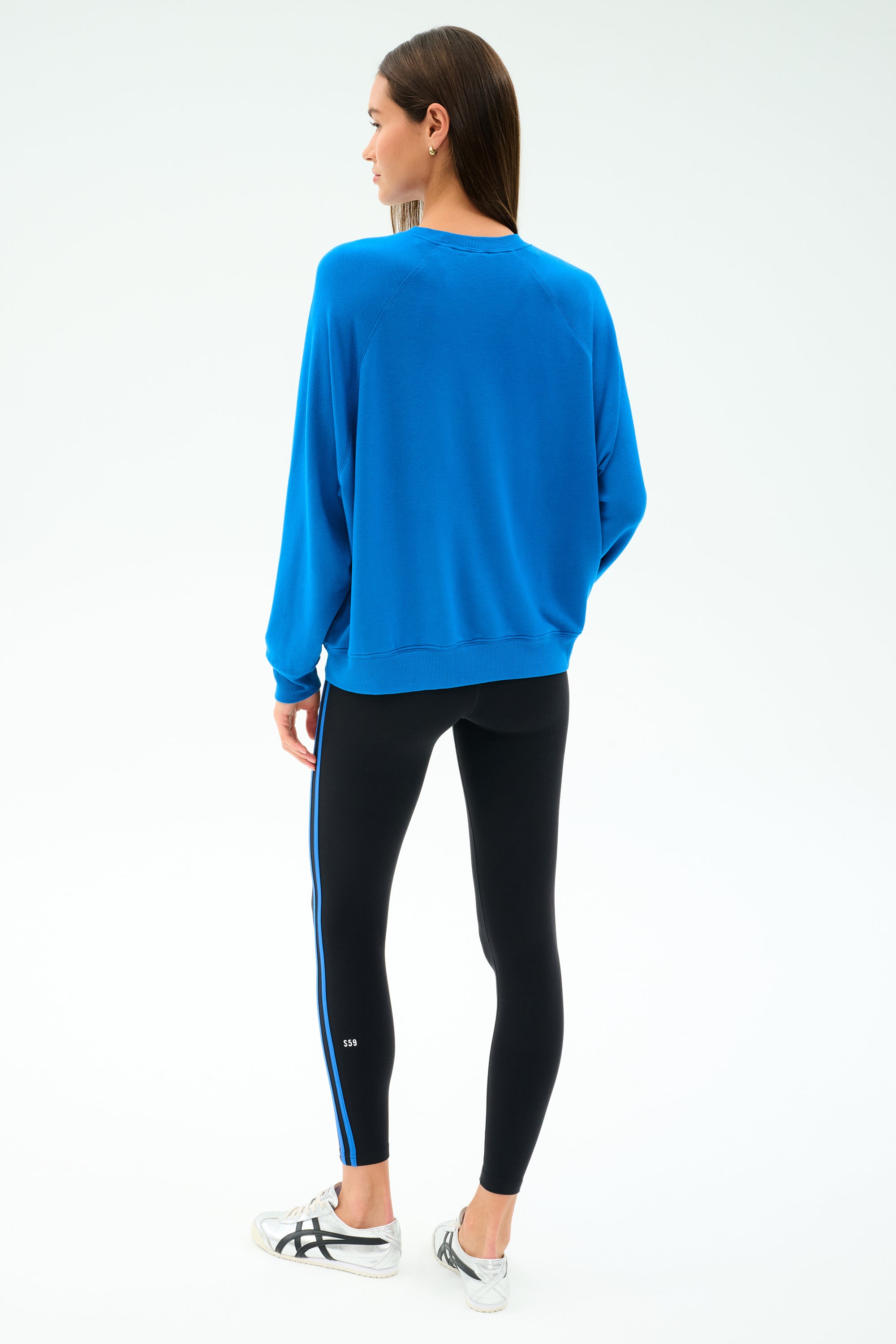 A woman stands facing away, wearing the SPLITS59 Andie Oversize Fleece Sweatshirt in Classic Blue with black leggings featuring a blue stripe, paired with white sneakers against a plain white background.