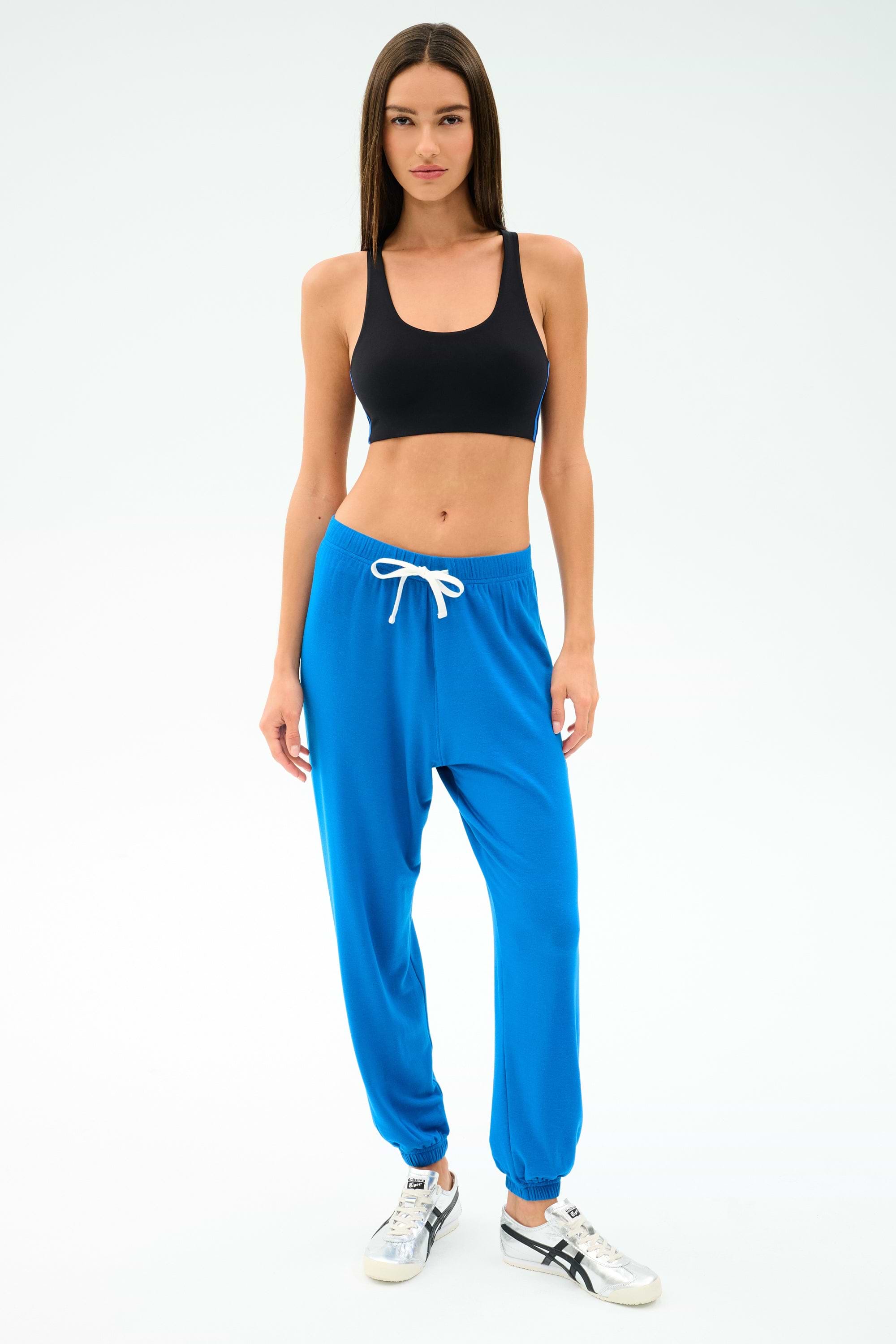 A woman faces forward against a white background, wearing a black sports bra, white sneakers, and SPLITS59 Andie Oversized Fleece Sweatpant in Classic Blue with a white drawstring.