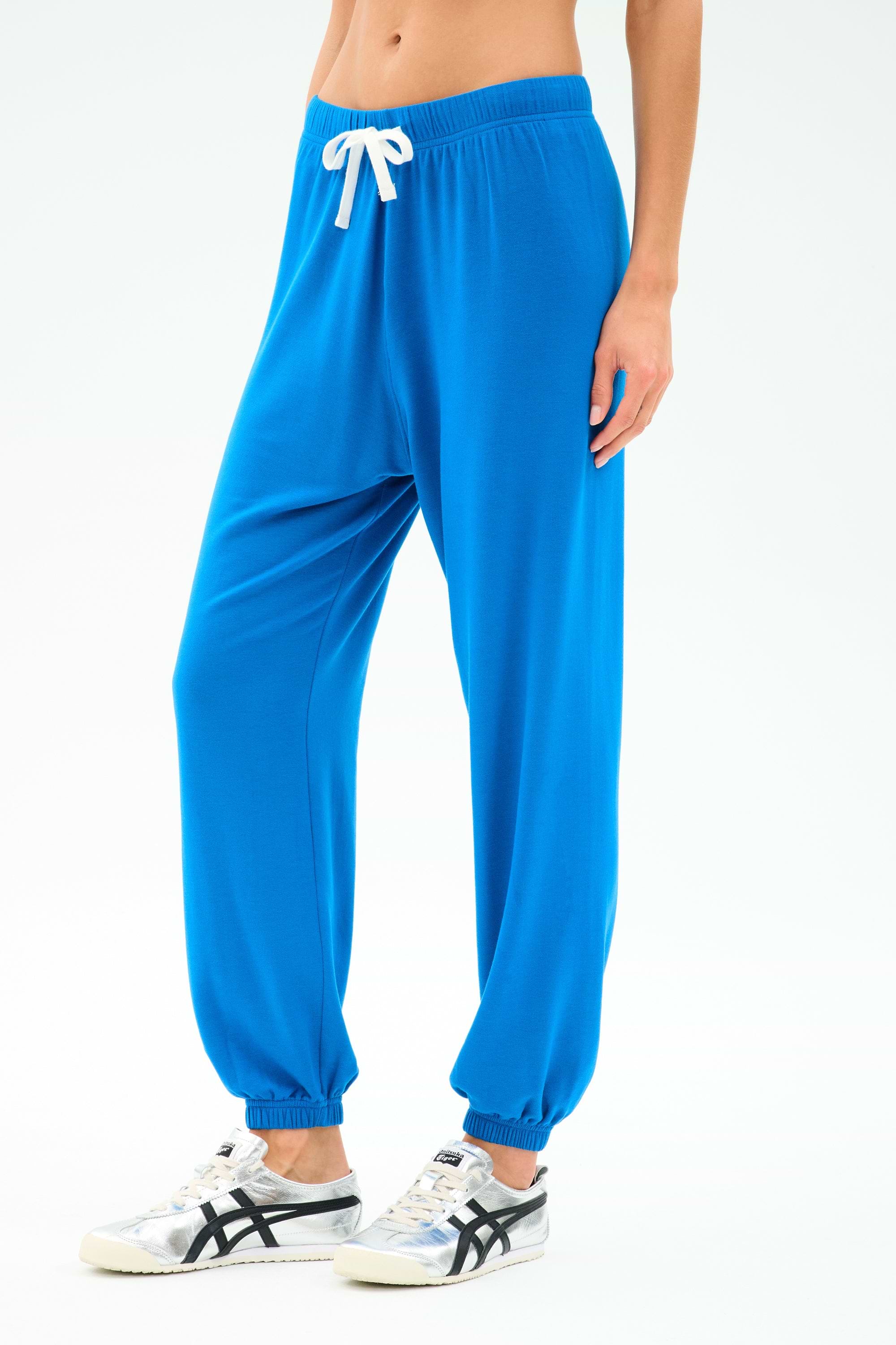 Person wearing SPLITS59 Andie Oversized Fleece Sweatpant in Classic Blue with a white drawstring and white sneakers with black details, standing against a plain light background.