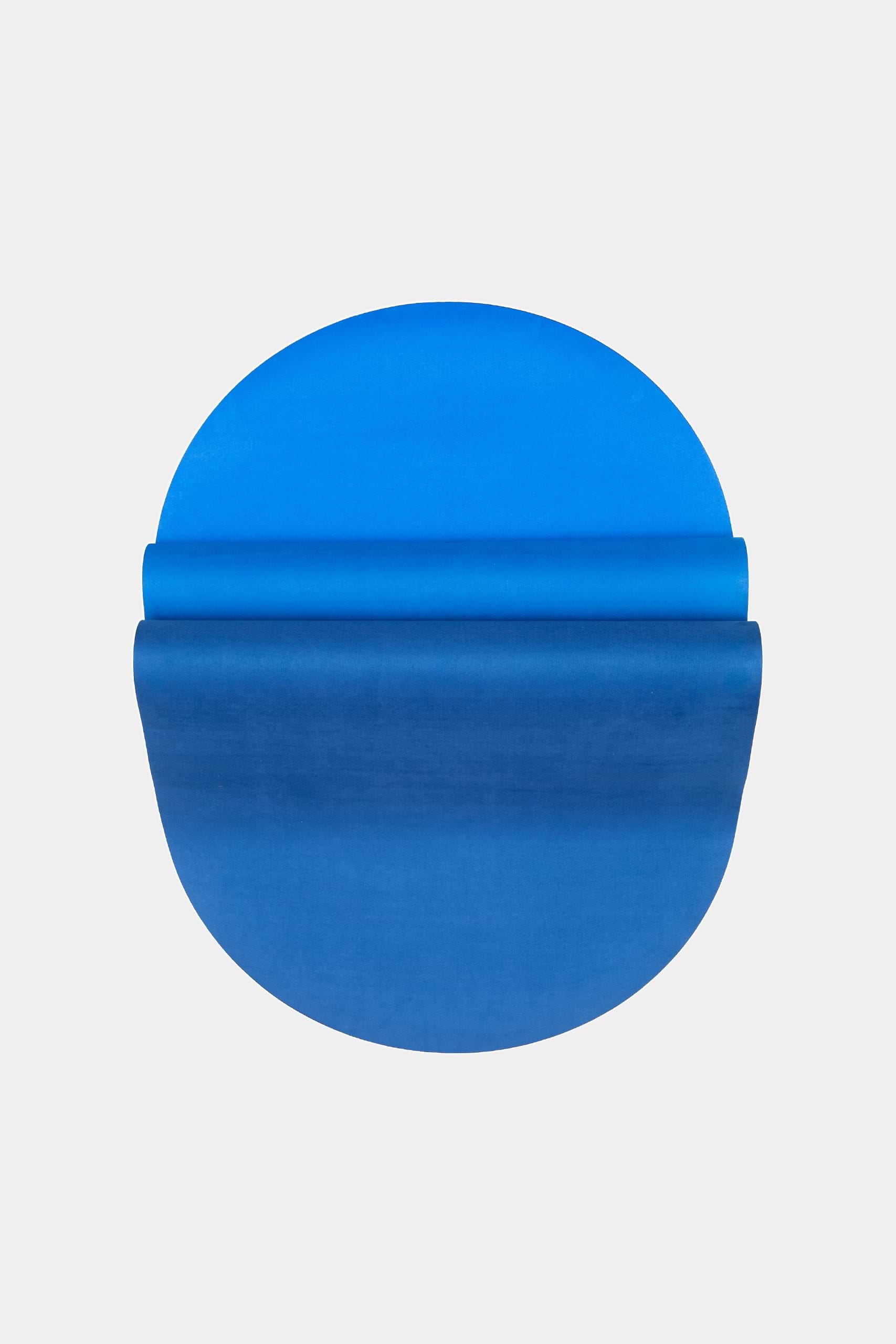 A round blue object with a gradient effect, resembling The Skye Mat by SPLITS59—an eco-rubber yoga mat—partially unrolled or folded over itself on a plain light gray background.