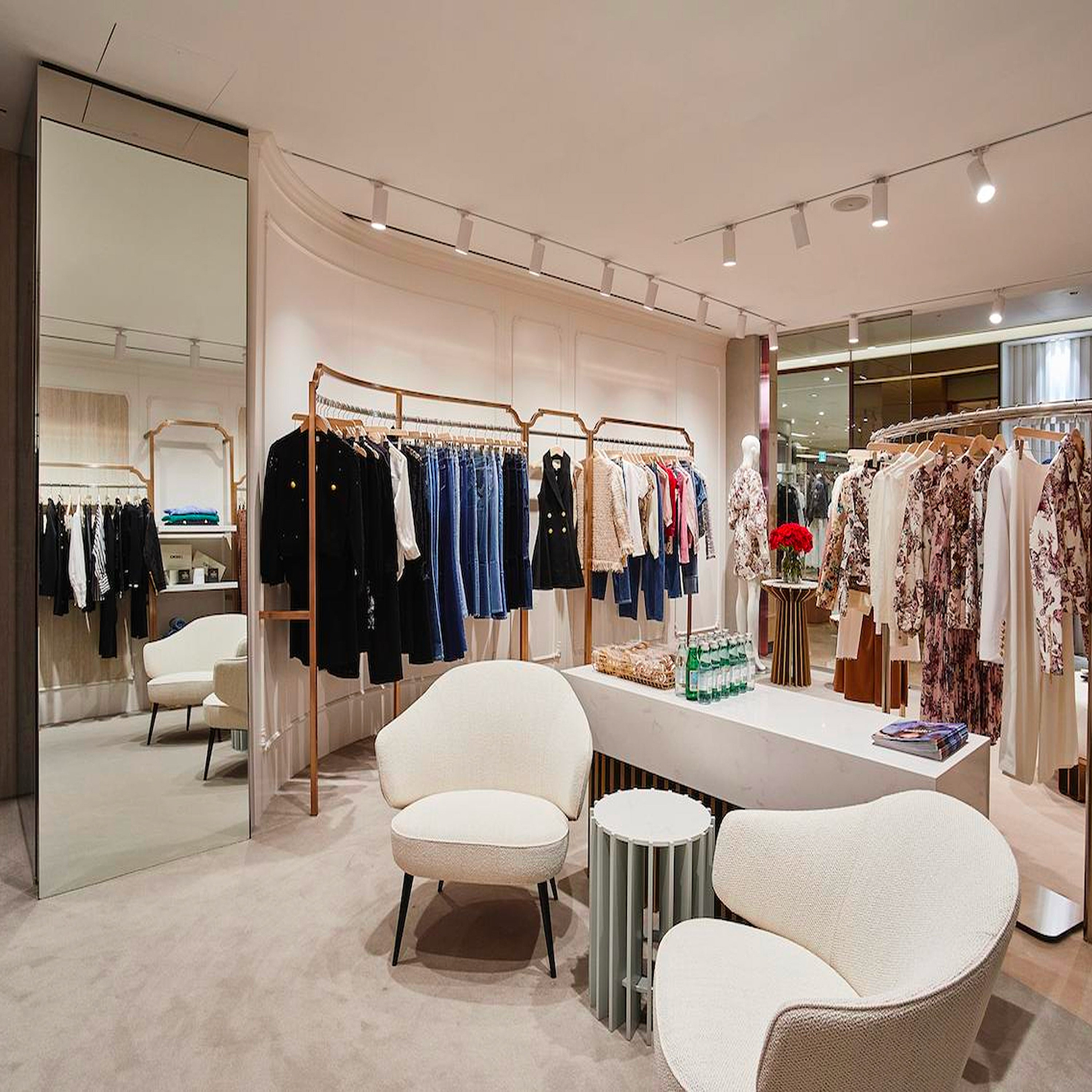Una boutique di abbigliamento elegante con espositori di jeans, giacche e abiti floreali, un grande specchio, due moderne sedie bianche, un tavolino e un tavolo espositivo con fiori e bottiglie d'acqua.