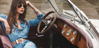 A woman with long brown hair lounges on a tan leather sofa, wearing a white sleeveless top, blue flared jeans, brown belt, and brown boots, with dark wood furniture in the background.
