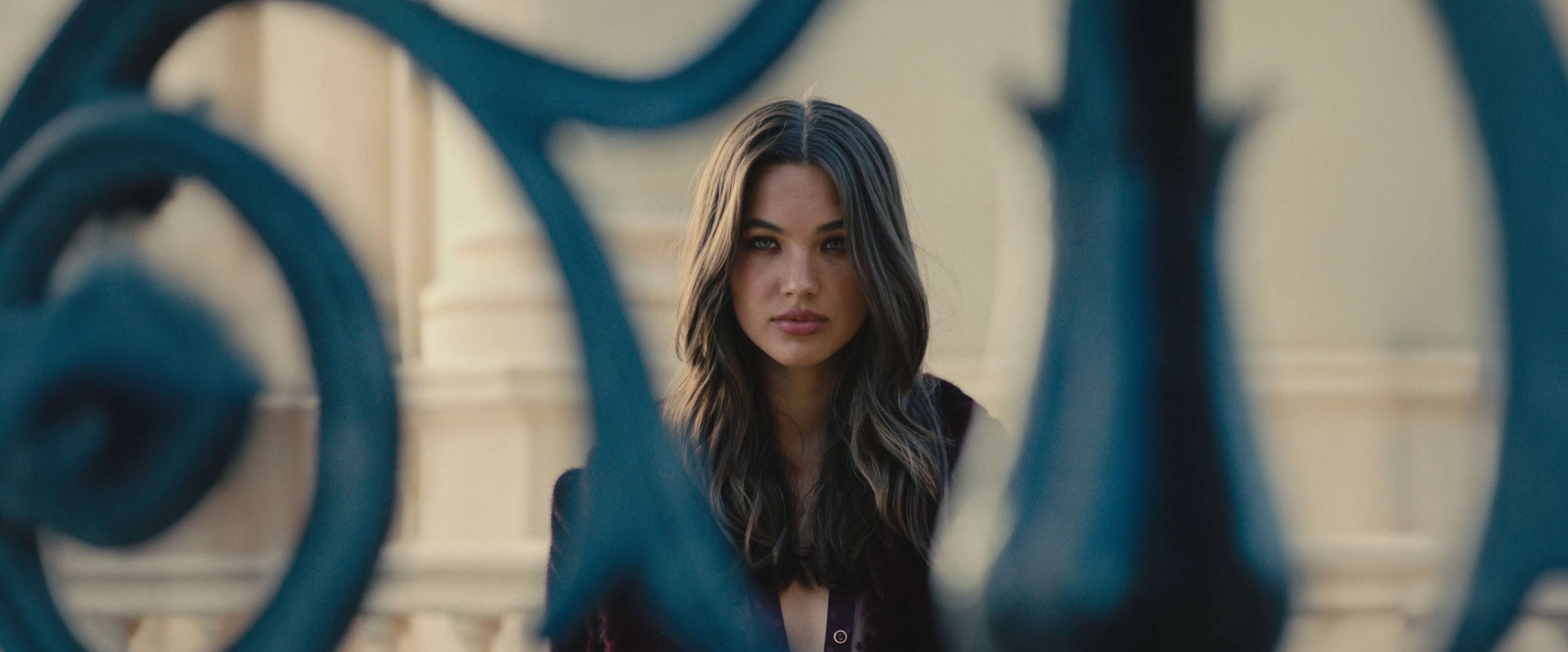 A woman with long, wavy hair stands behind ornate, blurred wrought iron bars, looking directly at the camera with a neutral expression. The background is softly lit and architectural.
