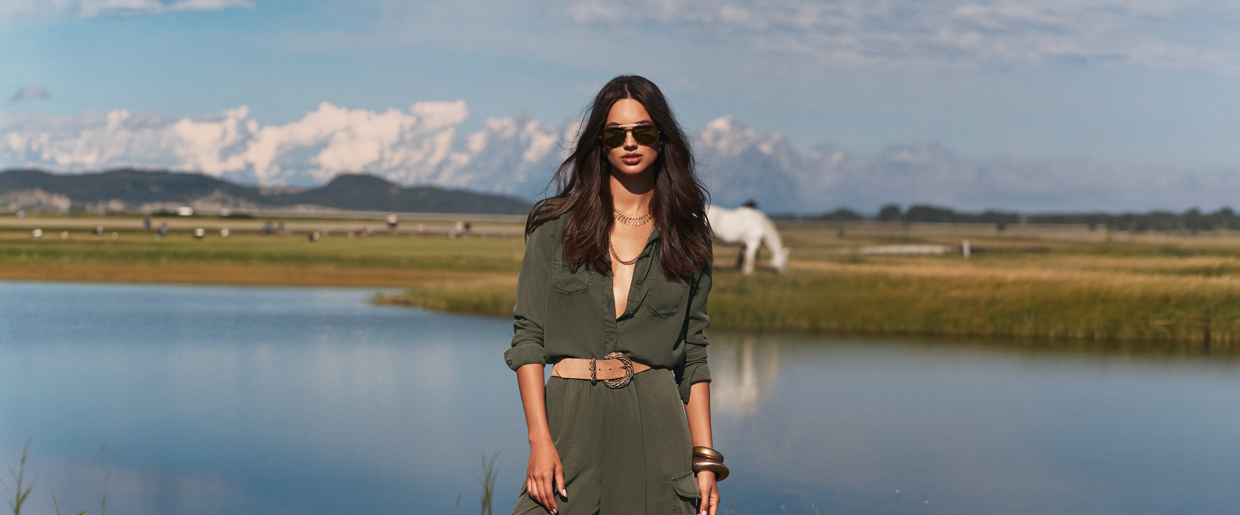 A woman in a green jumpsuit and sunglasses stands by a lake with mountains, grass, and a white horse in the background under a partly cloudy sky.