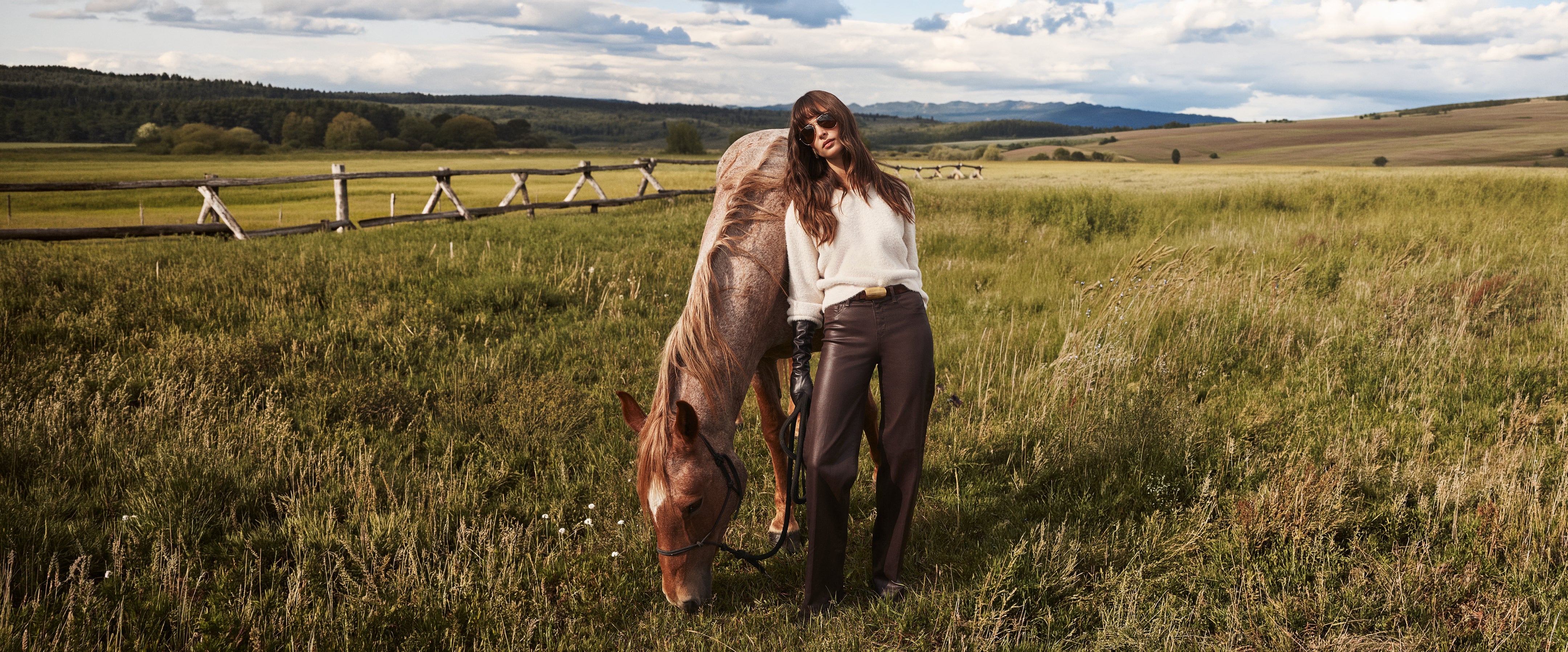 A woman in sunglasses and brown pants stands in a grassy field with a horse grazing beside her. A wooden fence and rolling hills are visible in the background under a partly cloudy sky.