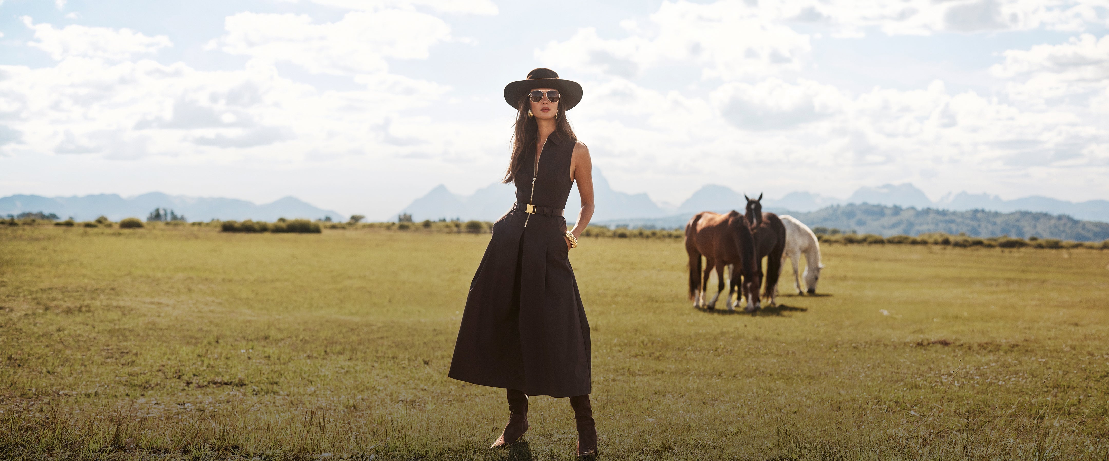 A woman wearing sunglasses and a strapless dress stands in the middle of a grassy, tree-lined path in a forest, hands in her pockets, looking confidently at the camera.
