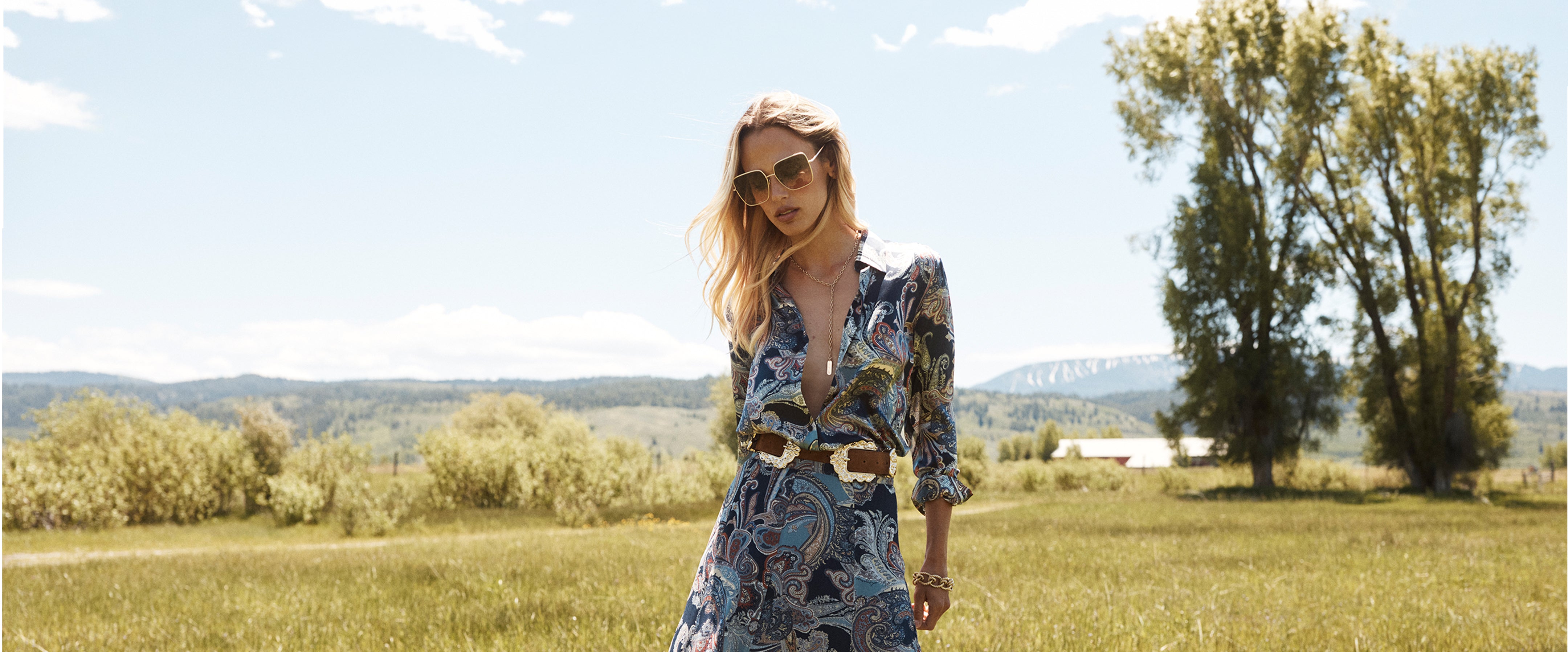 A woman in sunglasses and a patterned dress stands in a sunny field with trees, hills, and a blue sky in the background.
