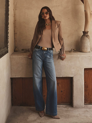 A woman with an hourglass figure wears the Vashti Fringed Leather Blazer, a beige top, blue wide-leg jeans, taupe heels, sunglasses, and gold jewelry as she leans against a rustic wall in a neutrally decorated room.