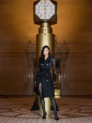 A woman poses confidently in front of a large gold clock. She wears a black sleeveless trench coat over a sheer black lace bodysuit and black heels. The setting is a grand indoor space with ornate architectural details.