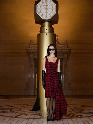 A woman in the L'AGENCE Aya Tweed Midi Dress from Fall 25 NYFW, with black gloves, heels, and sunglasses, stands before a grand gold clock in an ornate building, holding her matching coat.