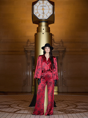A woman in a red floral-patterned suit and black hat stands in front of a large gold clock inside an elegant building with ornate architectural details. She wears black gloves and has long dark hair.