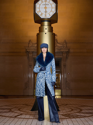 A person wearing a blue patterned coat with a fur collar, black gloves, leather pants, and a newsboy cap stands in front of a large gold clock on a column inside a grand, ornate hall.