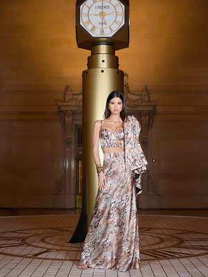 A woman in a brown and beige patterned two-piece outfit stands confidently in front of a large gold clock inside an elegant, ornate building with a tiled floor.
