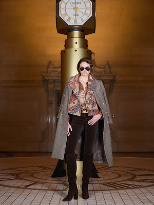 A woman poses confidently under a grand gold clock, wearing L'AGENCE Stevie Velvet Jeans with a patterned jacket, sunglasses, and a coffee bean brown coat in an elegant hall with tiled floors and ornate decor.