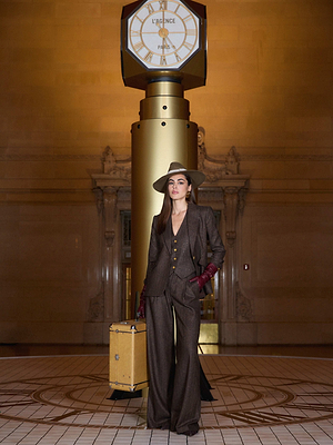 A stylish woman in L'AGENCE Allanah Pinstriped Trousers, a hat, and red gloves stands before a vintage clock, holding a yellow suitcase in an elegant train station.