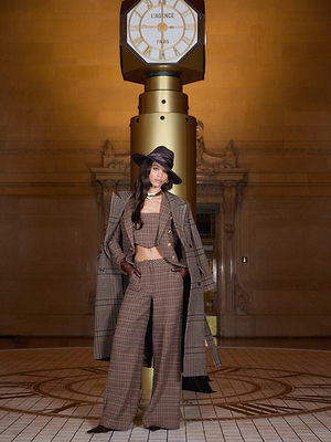 A woman in the Tiffany Corset Top by L'AGENCE poses confidently before a large gold clock, set in an elegant building with high ceilings and ornate details.