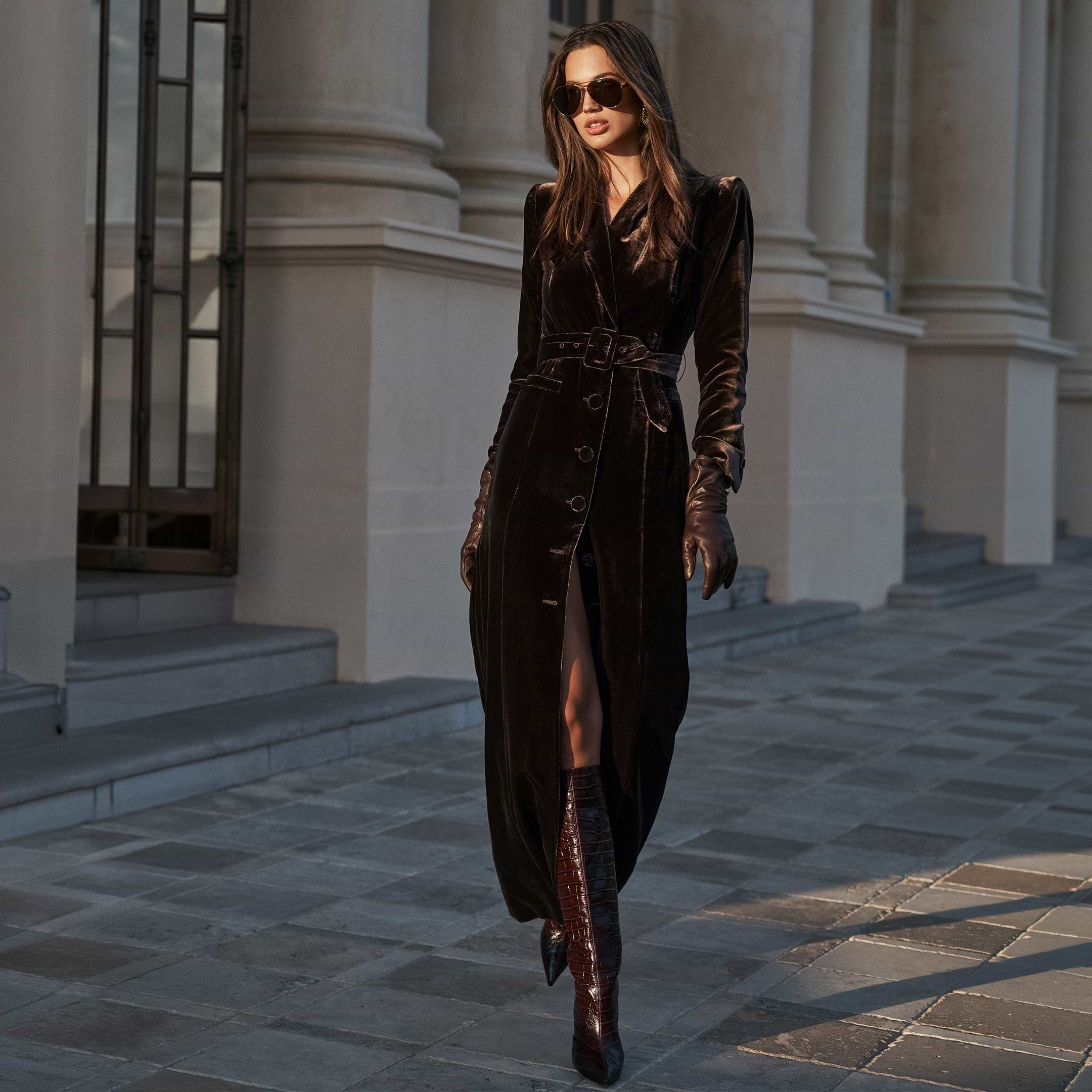 A woman wears a long, buttoned Raini Velvet Maxi Dress with a shawl collar, a belt, matching gloves, and knee-high boots, walking past a building with stone columns.