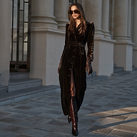 A woman wears a long, buttoned Raini Velvet Maxi Dress with a shawl collar, a belt, matching gloves, and knee-high boots, walking past a building with stone columns.