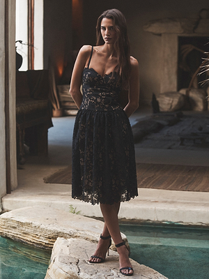 A woman in the Flyn Lace Midi Dress by L'AGENCE Sale stands by an indoor pool, hands behind her back. The black fit-and-flare dress features floral eyelet lace, and the rustic setting is filled with neutral tones and natural light.