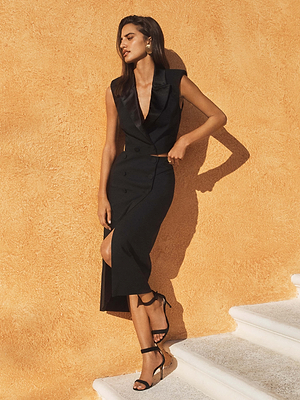 Wearing the L'AGENCE Hollis Vest, a woman stands on white stairs against a textured orange wall, posing in strappy heels with one hand on her hip and her head slightly tilted.