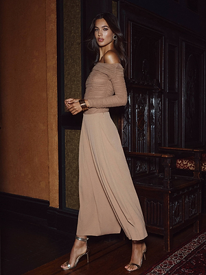 A woman poses indoors wearing the L'AGENCE Novalynn Cropped Pleated Pant with a tan off-the-shoulder top, gold bangles, and metallic heels. The scene includes dark wood paneling and ornate furniture.