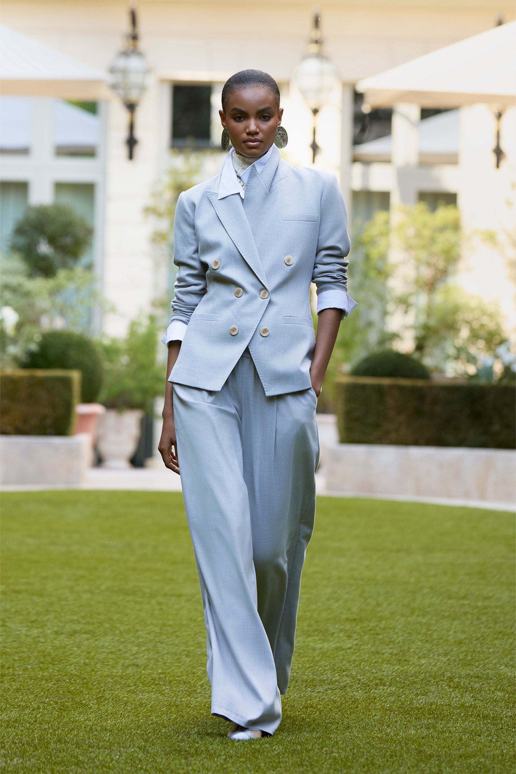A model walks outdoors on grass wearing a light blue double-breasted blazer and wide-leg trousers, hands in pockets. The scene features a building, umbrellas, greenery, and a modern fashion vibe.