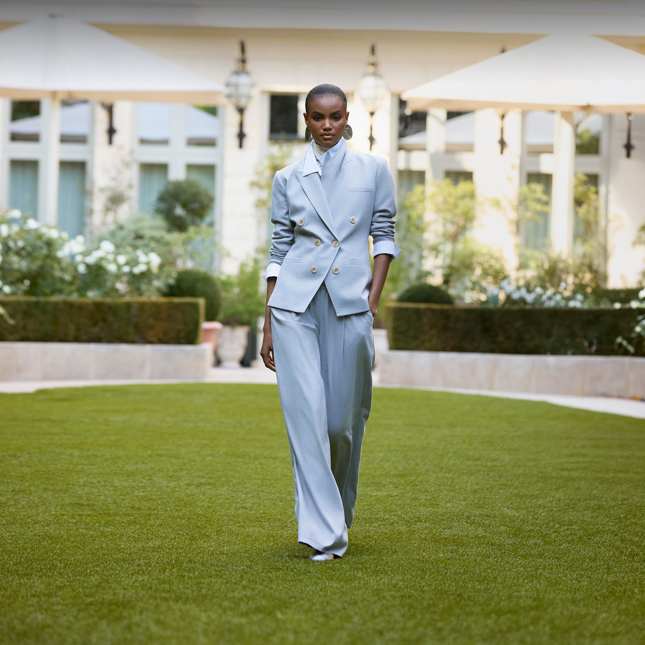 A model walks on grass in a light blue suit featuring the Kyomi Double-Breasted Blazer and wide-leg pants during a garden fashion presentation inspired by Paris Fashion Week Spring 2027.