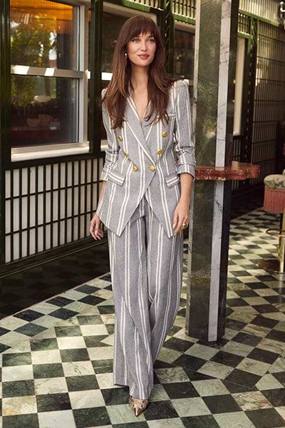 A woman with long brown hair wearing a gray pinstriped double-breasted suit and metallic pointed shoes stands on a green and white checkered floor in a stylish, sunlit room with patterned walls and large windows.