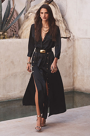 A woman with long brown hair wears a black belted dress with gold buttons and high slits, layered gold necklaces, bracelets, and metallic high heels, walking near a stone wall and indoor pool with plants.