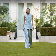 A model walks on a green lawn in front of a white building, wearing a light blue sleeveless belted vest and matching wide-leg trousers.