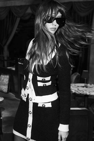 A woman with long hair wearing dark sunglasses and a black-and-white buttoned dress stands indoors, looking to the side as her hair swings, in a black-and-white photo with a stylish, elegant atmosphere.