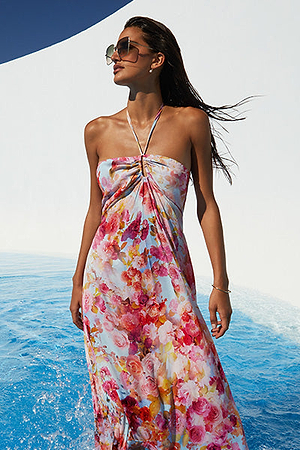 A woman wearing sunglasses and a colorful floral halter-neck dress stands in shallow blue water, with wet hair and a white wall and blue sky in the background.