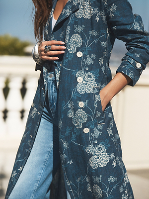 A person stands outdoors wearing a blue floral patterned trench coat over blue jeans, with one hand in the coat pocket and the other adjusting the lapel. The background is bright and blurred.