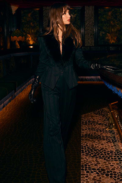 A woman in a black pinstripe suit with a fur collar and black leather gloves stands indoors, looking to the side. The setting has dim lighting and a patterned stone floor.