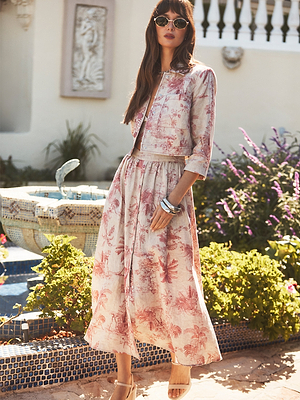 A woman in sunglasses stands in a garden by a fountain, wearing a light pink floral two-piece outfit with a cropped jacket and a long skirt, accessorized with bracelets and nude heels.