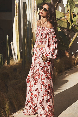 A woman in sunglasses wears a matching off-shoulder top and wide-leg pants with a red and white geometric pattern, standing outdoors near cacti and dry grass in bright sunlight.
