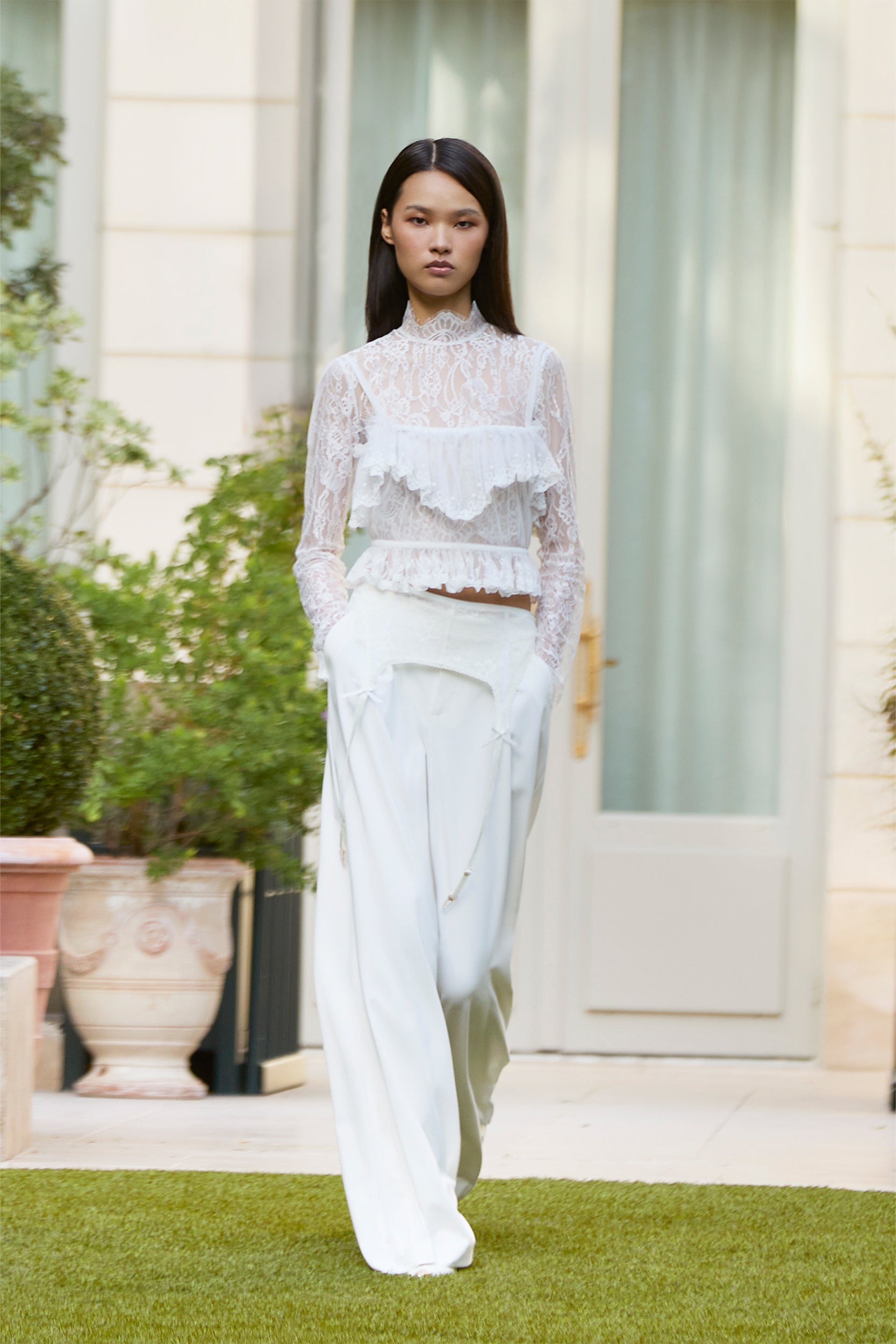 A woman strolls on grass in a white lace long-sleeve blouse and high-waisted white trousers, hands in her pockets. Potted plants and a glass door form the background.