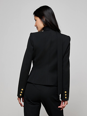 A woman with long dark hair models the L'AGENCE Romy Tie Neck Blazer, featuring gold buttons and paired with matching black pants, as she stands facing away from the camera against a light gray background.