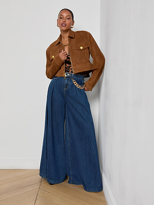 A woman stands against a white wall wearing the Micah Oversized Suede Jacket, a patterned top, and high-waisted wide-leg blue jeans with an oversized fit, styled with a gold chain belt, gold jewelry, and hoop earrings.