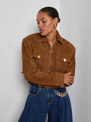 A woman stands against a white wall wearing the Micah Oversized Suede Jacket in brown suede with gold buttons, paired with wide-leg blue jeans and gold hoop earrings. Her hair is pulled back and she looks down with arms crossed.
