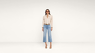 A woman wearing sunglasses, a beige jacket, light blue cropped jeans, and heeled sandals stands against a plain white background.